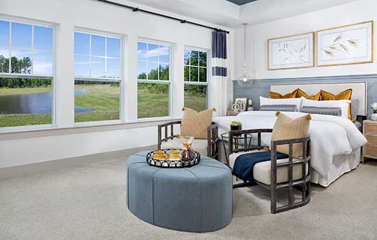 A decorated model home Owner’s Retreat with three large windows open and looking out over a scenic lake surrounded by trees. Two cozy chairs and a small table are situated near the foot of the bed.