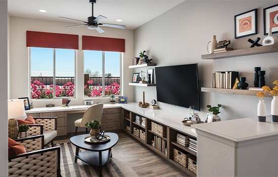 This David Weekley model home’s study features soft red window shams, woven textile shairs, and a rounded triangular coffee table as focal points of the room’s transitional design style, with a row of bright pink flowers visible through the large windows.