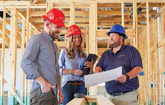 Three people wearing hardhats are looking at the same piece of paper. They’re inside a new home currently under construction, and the treated lumber of the frame of the home can be see around them.