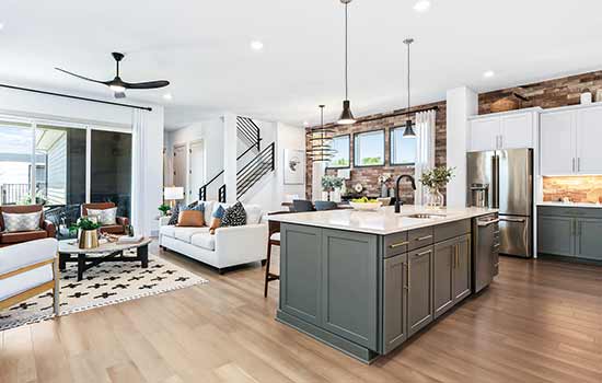 The interior of a model home. A quartz-topped kitchen island and green-and-white kitchen cabinets are set to the right. To the left is an open living space with mixed white-and-brown furniture and big windows overlooking the backyard. Wood floors flow through all these spaces. A stone-accented dining space and a staircase with black hand rails can be seen in the background.