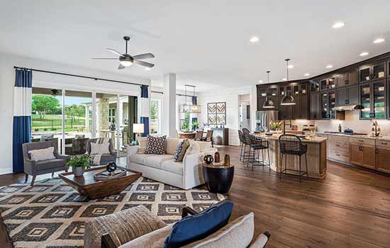 The primary living spaces inside a model home. Gray, brown and white balance in the family room on the left. Two-tone wood cabinets accent the spacious kitchen to the right. Large, energy-efficient windows allow light to enter without allowing heat to intrude, and provide a beautiful view of the greenery behind the home.