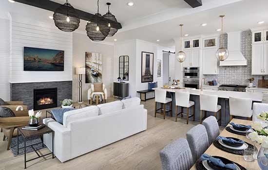 The interior living spaces of a model home. A fireplace creates a focal point in the white, blue and brown family room with black rafters and light fixtures above. Places are set for eight at the dining table to the right. White chairs, countertops, cabinets and backspash create a unified depth of style in the open kitchen at the back of this photo.