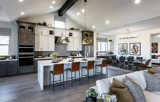 The open living spaces of a model home with white and gray details in the kitchen, a dark wood beam spanning the ceiling and an elegantly appointed dining space in background.