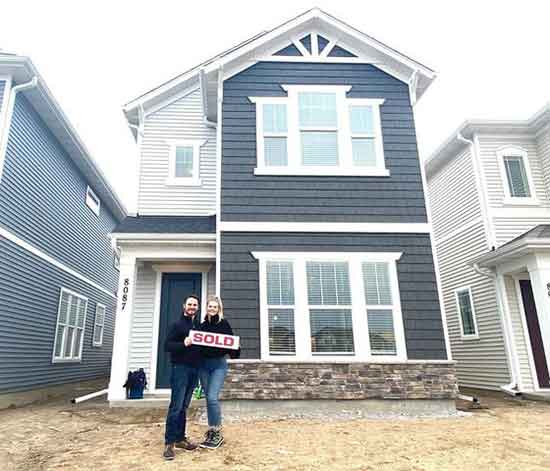 new homeowner couple standing in front of their newly constructed home holding a SOLD sign