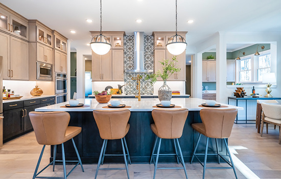 A model home kitchen photo showing bar stools at the kitchen island, pendant lighting, a classy backsplash and the expansiveness of the home beyond the kitchen.