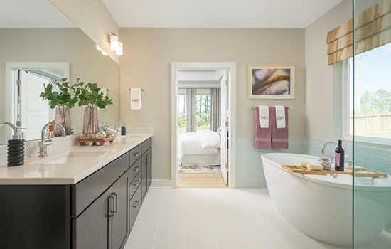 This David Weekley model home’s Owner’s Bath shows dark cabinetry and white quartz countertops on the left, a glass-enclosed shower and free-standing tub on the right, and the Owner’s Retreat just outside the door.