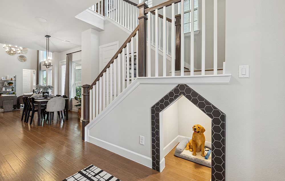 A David Weekley model home’s interior, with hardwood floors leading to the dining and family areas to the left, and hardwood accents ascending the staircase, where a tile-lined opening reveals a finished space under the stairs, where a prop Labrador is situated on a dog bed.
