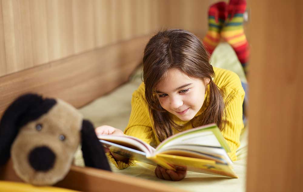 A young girl is reading a children’s book with a stuffed animal nearby on a small bed built into the space under the stairs.
