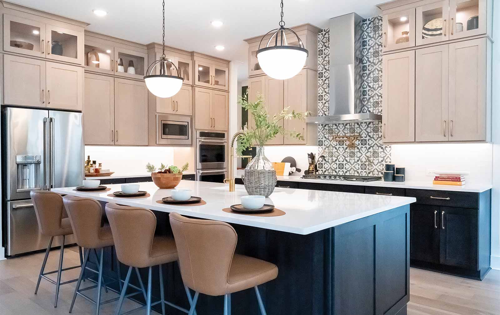 Black lower cabinets are topped by white quartz countertops, which give way to tan cabinetry that matches the tan stools pulled up to the kitchen island in the foreground. Upper cabinets feature glass panes, showcasing a variety of pieces of decorative pottery within.
