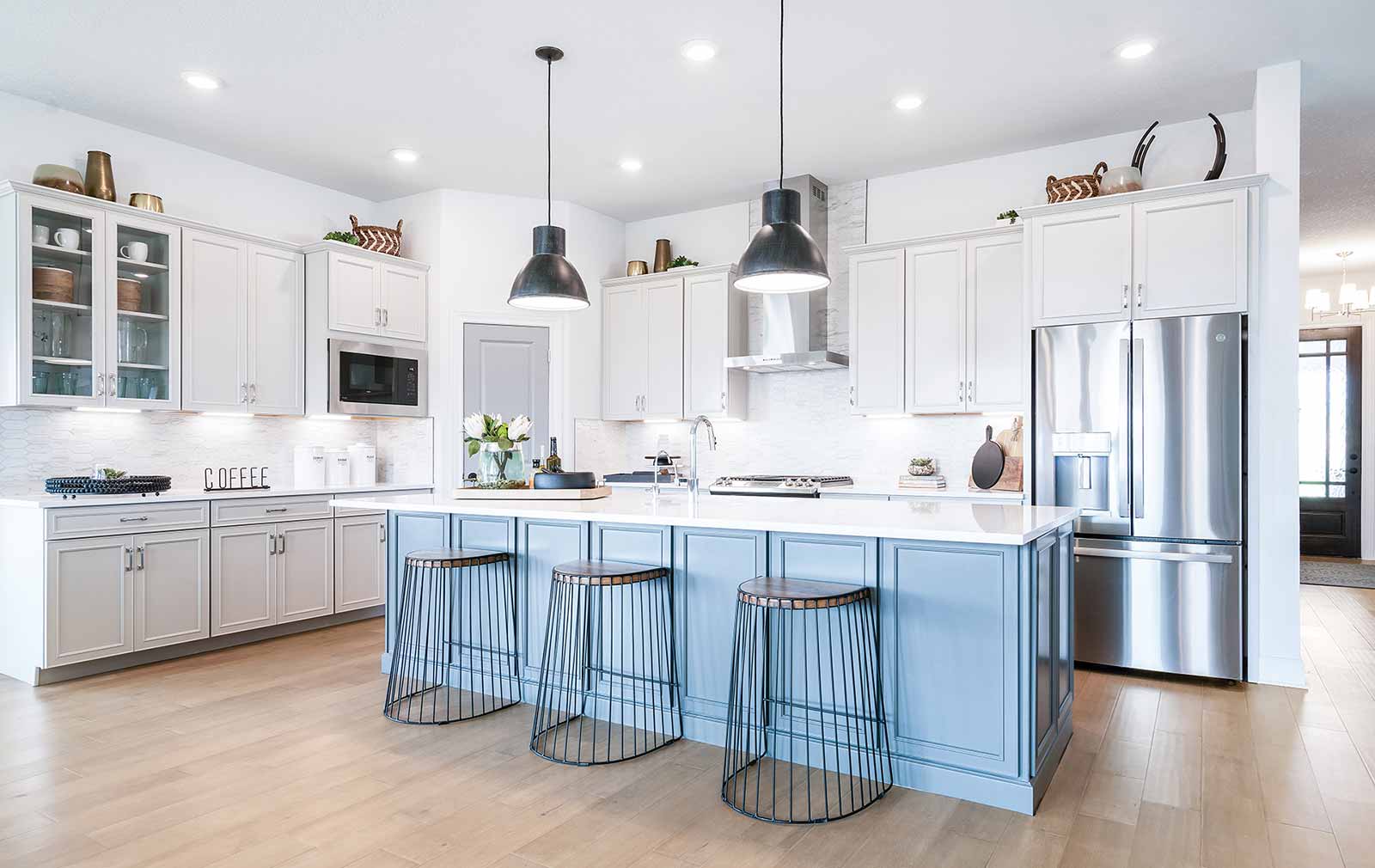 Rod iron spoke stools are pulled up to the soft blue island, all surrounded by white cabinets and stainless steel appliances. The white backsplash helps add height to the countertops by reflecting the natural and recessed light.