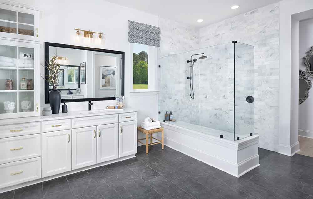 The Owner’s Bath of a David Weekley model home with gray stone tile floors, white cabinetry with a built-in cupboard beside the vanity and sink, white quartz countertops, and a glass partition separates the Super Shower’s white tile from the rest of the Owner’s Retreat.