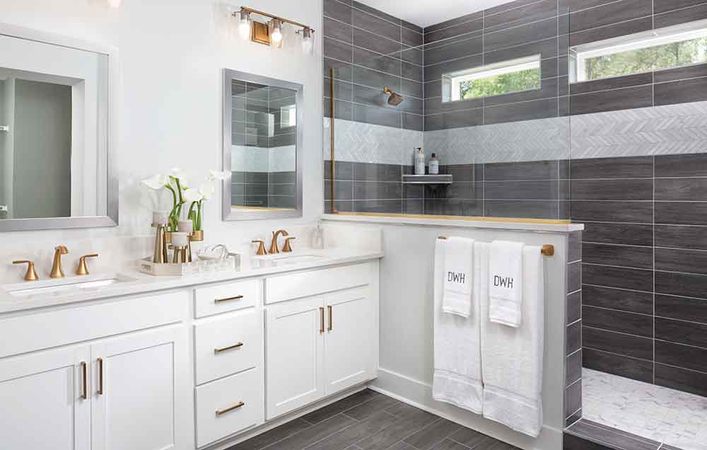 The Owner’s Bath of a David Weekley model home with light gray stone tile floors, white cabinetry, striated quartz countertops, cobbled stone and gray tile walls, and a large, free-standing tub in front of the glass partition separating the Super Shower from the rest of the space.
