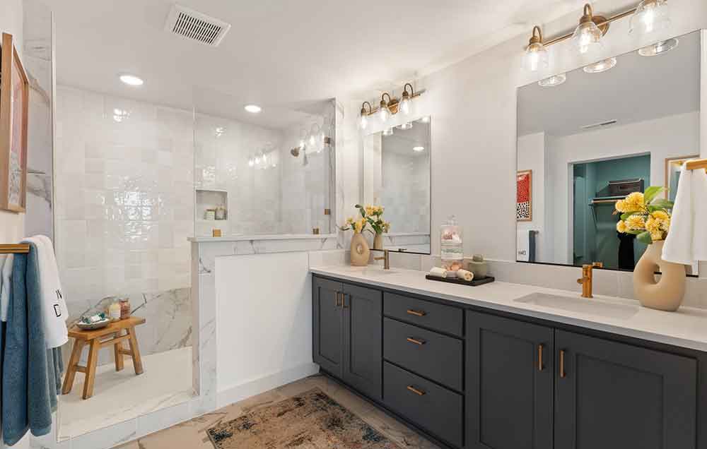 The Owner’s Bath of a David Weekley model home with stone tile floors, gray-blue cabinetry, white quartz countertops, and marble accents and subway tile in the Super Shower.