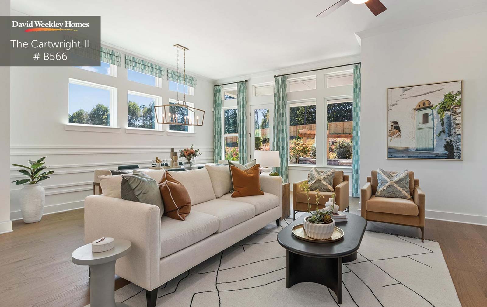 A living room in the foreground with white walls, furniture, and carpet, all touched with tan, brown, gold, green and wood accents along with natural textures. Green and white curtains frame the window view of the lush garden in the backyard. 