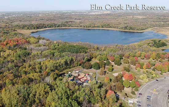 An aerial view of a lush, forested expanse of park land surrounded a scenic lake with a small parking lot to the side providing access to a playground and walking trails nestled among the trees.